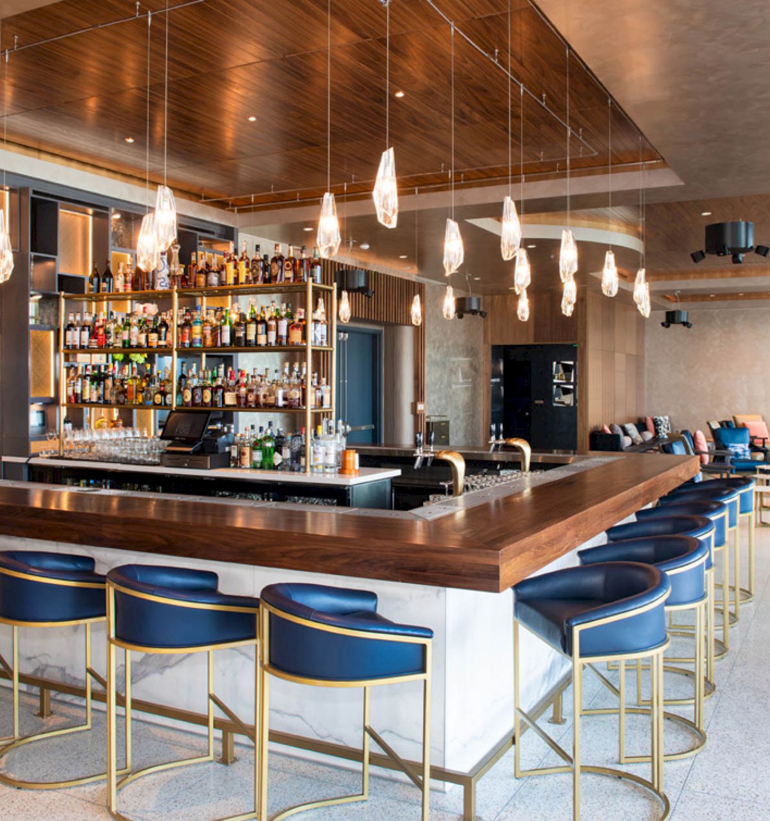 A modern bar with blue stools, hanging lights, and a stocked shelf of bottles, set in a spacious, stylish lounge area.