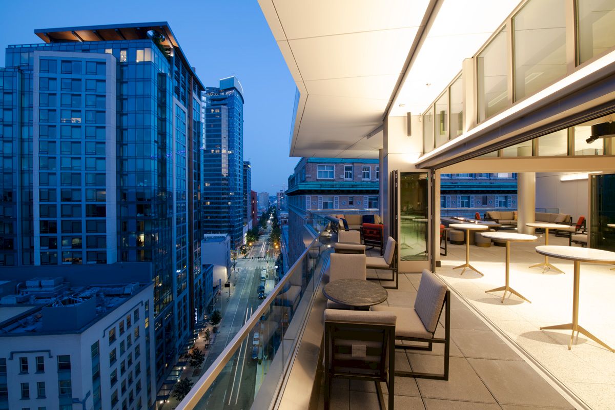 A modern city view from a balcony at dusk, featuring glass buildings, outdoor seating, and warm interior lighting.
