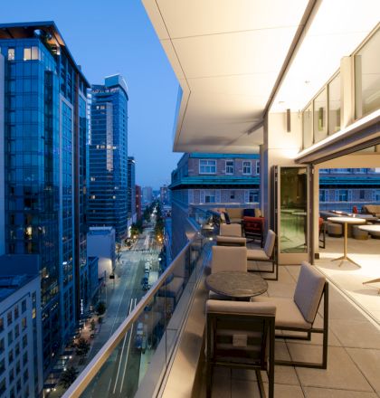 A modern balcony overlooking a cityscape with tall buildings at dusk, featuring seating and tables, warmly lit from inside.