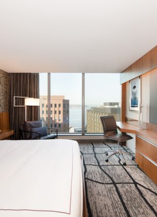 A modern hotel room features a large bed, desk, TV, and window with a city view, combining comfort with contemporary design.