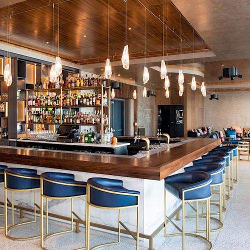 The image shows a modern bar with blue stools, hanging lights, and a well-stocked liquor shelf, surrounded by casual seating.