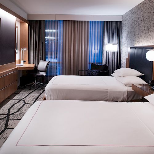 A modern hotel room with two beds, a TV, a desk with a chair, and patterned wallpaper, featuring soft lighting and a carpeted floor.