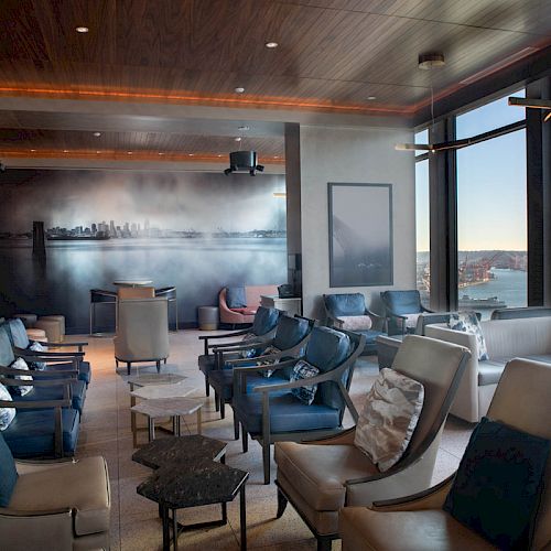 A modern lounge with leather chairs, large windows offering a waterfront view, and a mural on the wall.