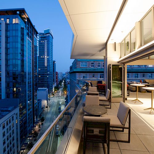 A modern cityscape at dusk with a view from a balcony, featuring tall buildings and a furnished outdoor seating area.