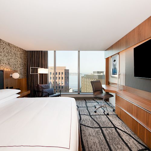 A modern hotel room with a king-size bed, large TV, desk, and chair overlooks a cityscape through a window.