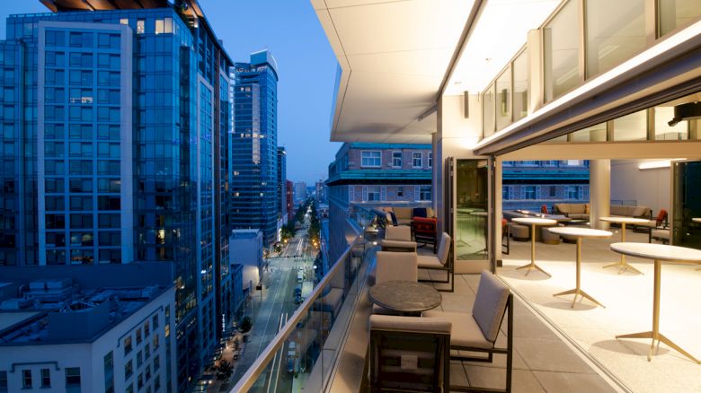 A modern balcony overlooking a city street lined with tall buildings, featuring outdoor seating and lighting as dusk falls.