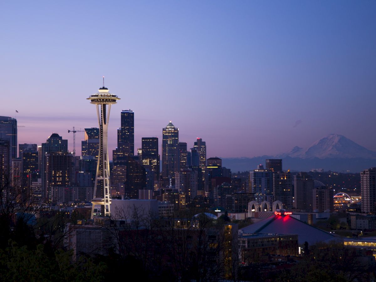 A city skyline at dusk with a prominent tower and a distant mountain, under a clear gradient sky.