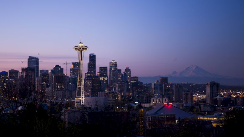 A city skyline at dusk with a prominent tower and a distant mountain, under a clear gradient sky.