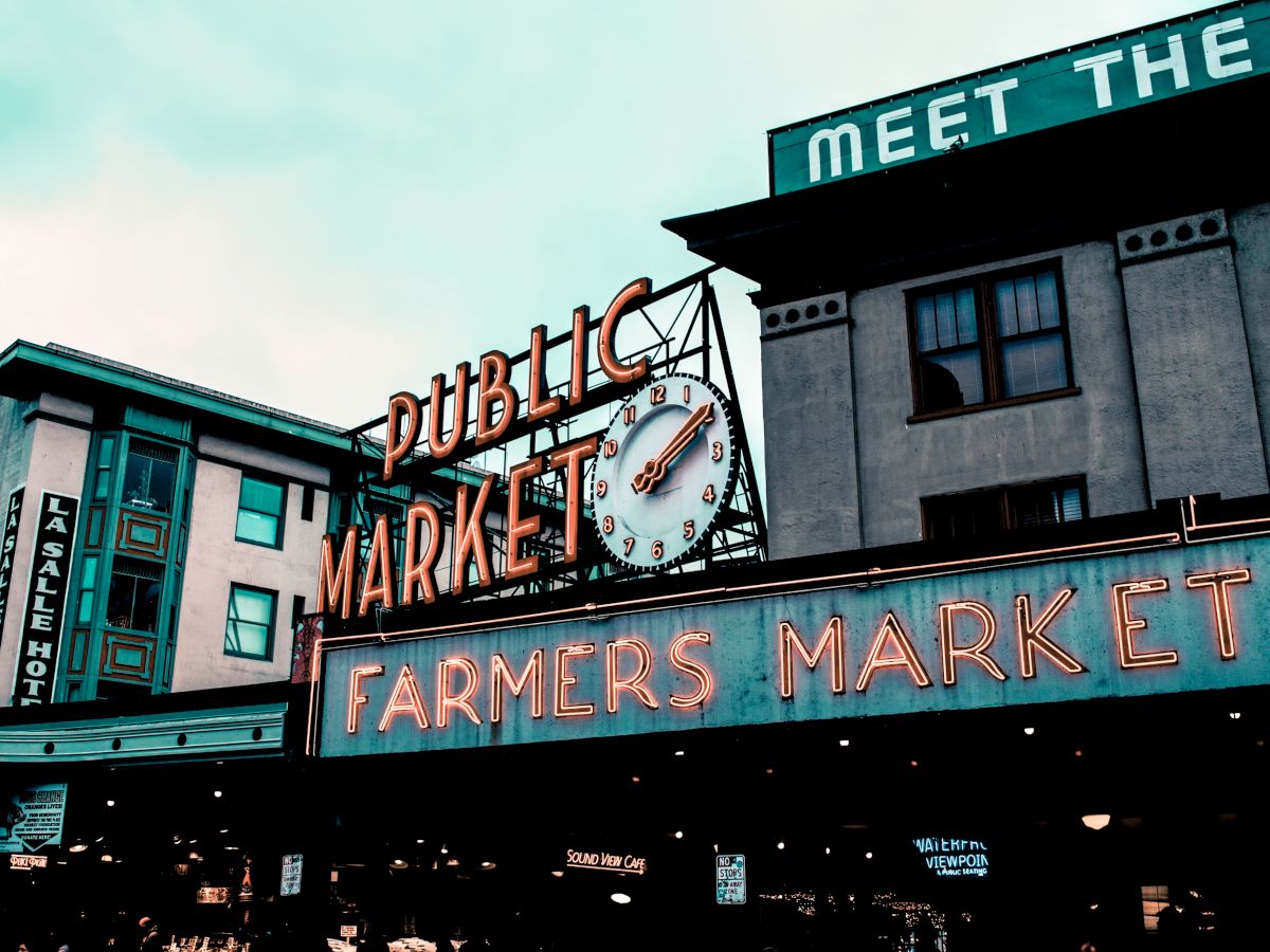A public market building with "PUBLIC MARKET" and "FARMERS MARKET" neon signs, featuring a clock. The city vibe is bustling and lively.