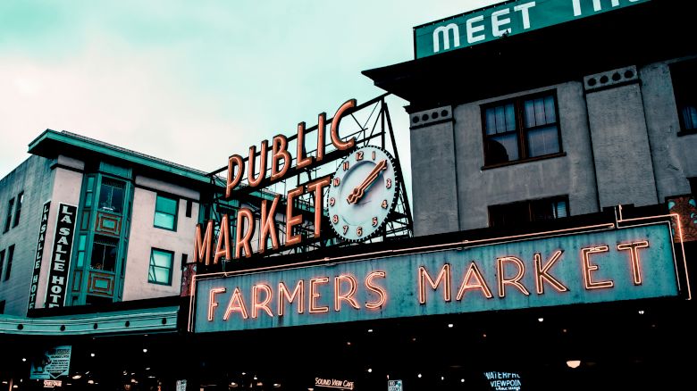 A public market building with "PUBLIC MARKET" and "FARMERS MARKET" neon signs, featuring a clock. The city vibe is bustling and lively.