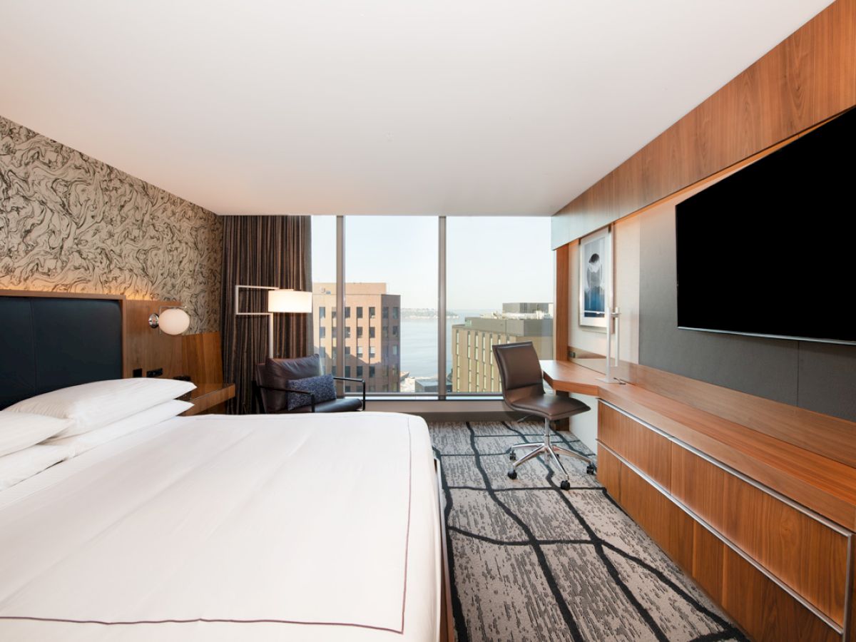 A modern hotel room with a large bed, wall-mounted TV, desk, and window showing a cityscape view.