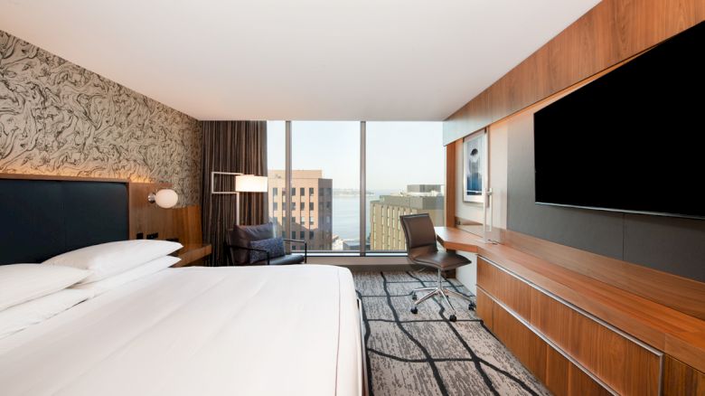 A modern hotel room with a large bed, wall-mounted TV, desk, and window showing a cityscape view.
