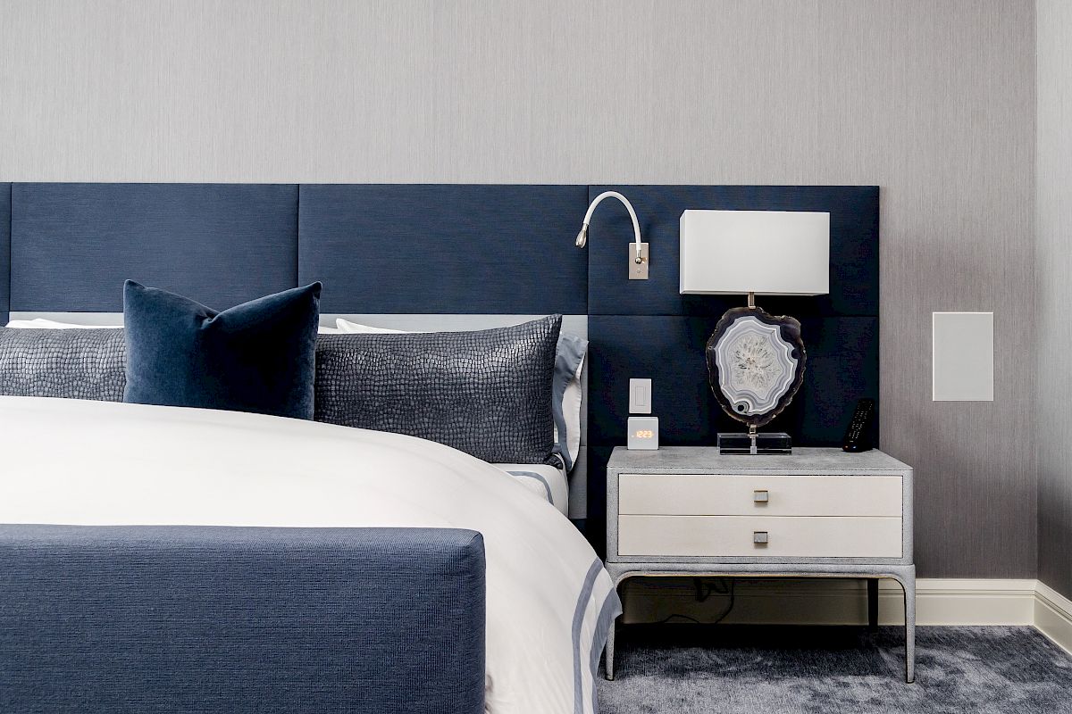 A modern bedroom features a navy bed, gray accents, a nightstand with a lamp, and a decorative object, all on a plush carpet.