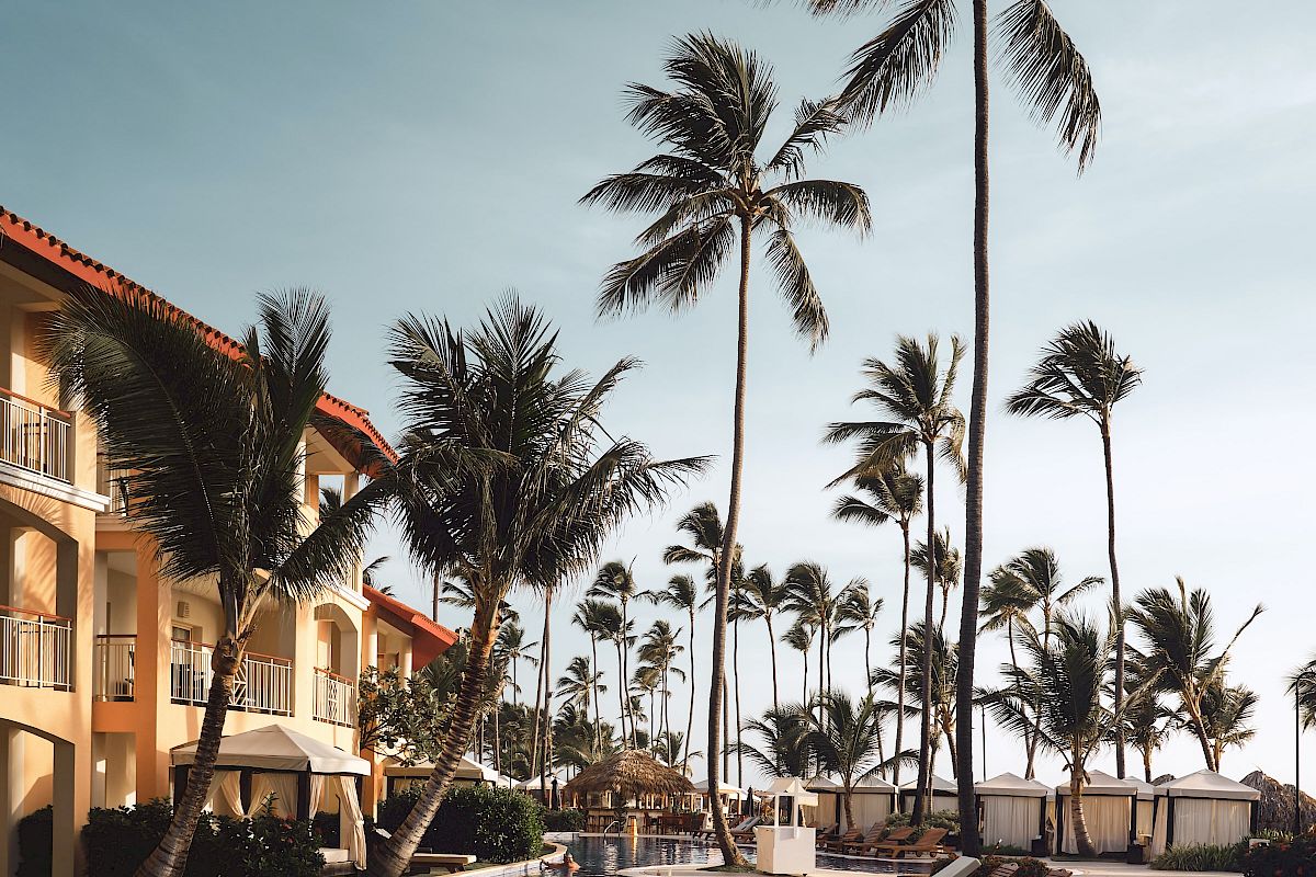 A scenic view of a resort with a swimming pool, lined with lounge chairs and surrounded by tall palm trees under a clear sky.