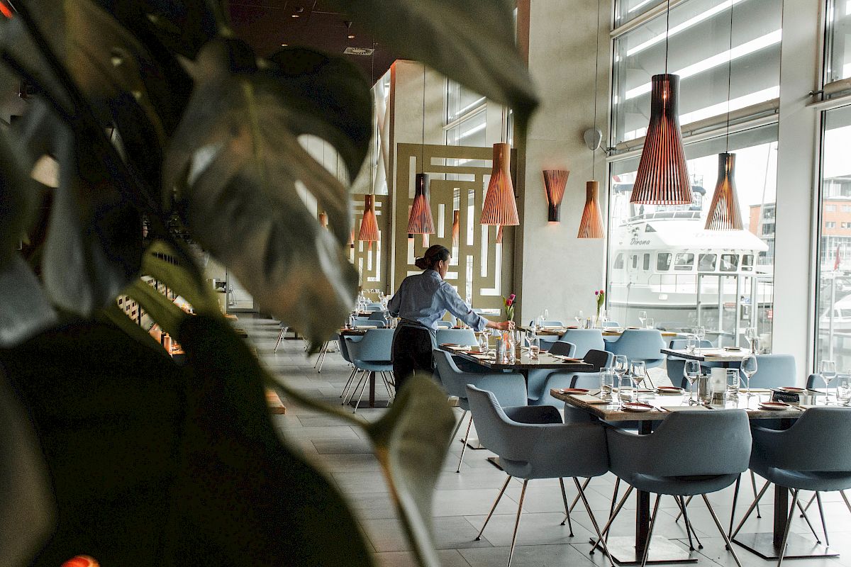 The image shows a modern restaurant with a person setting tables, stylish chairs, warm lighting, and large windows showing an urban view.