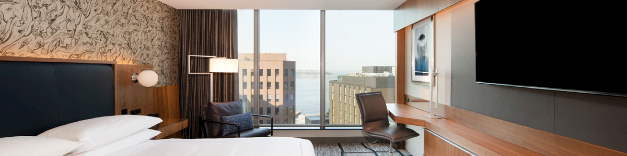A modern hotel room with a large bed, a TV on a wooden cabinet, a desk, and a city view through floor-to-ceiling windows.